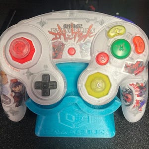 May include: A clear, custom-designed video game controller with red, green, yellow, and orange buttons. The controller has a black directional pad and a light blue stand. The controller has Japanese text and character graphics.