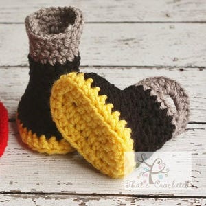 May include: A pair of crocheted baby booties in black, yellow and grey. The booties have a yellow sole and a black and grey upper.