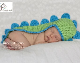 May include: A newborn baby wearing a green and blue crocheted dinosaur hat and matching tail. The baby is sleeping on a white blanket with a white and gray polka dot pattern.