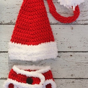 May include: Red and white crocheted Santa hat and diaper cover for a baby. The hat has a long, curled tail and a white pom-pom. The diaper cover has a white trim and a black button.