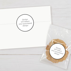 May include: A white envelope and a clear plastic bag containing a cookie. Both items have a round sticker with the text "Sticker will reflect your purchased design". The background is a light wood surface.