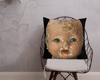 Puede incluir: Una almohada negra con una cara de muñeca de porcelana agrietada y espeluznante impresa en ella. La muñeca tiene ojos verdes y una boca abierta y agrietada.