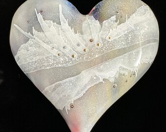 Puede incluir: Un corazón de vidrio blanco con un diseño de mármol en espiral y motas doradas. El corazón tiene un ligero tono rosado en la parte inferior.