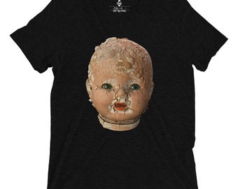 Puede incluir: Una camiseta negra con la cabeza de una muñeca de porcelana vintage impresa en la parte delantera. La cara de la muñeca está agrietada y astillada, lo que le da un aspecto espeluznante y misterioso.