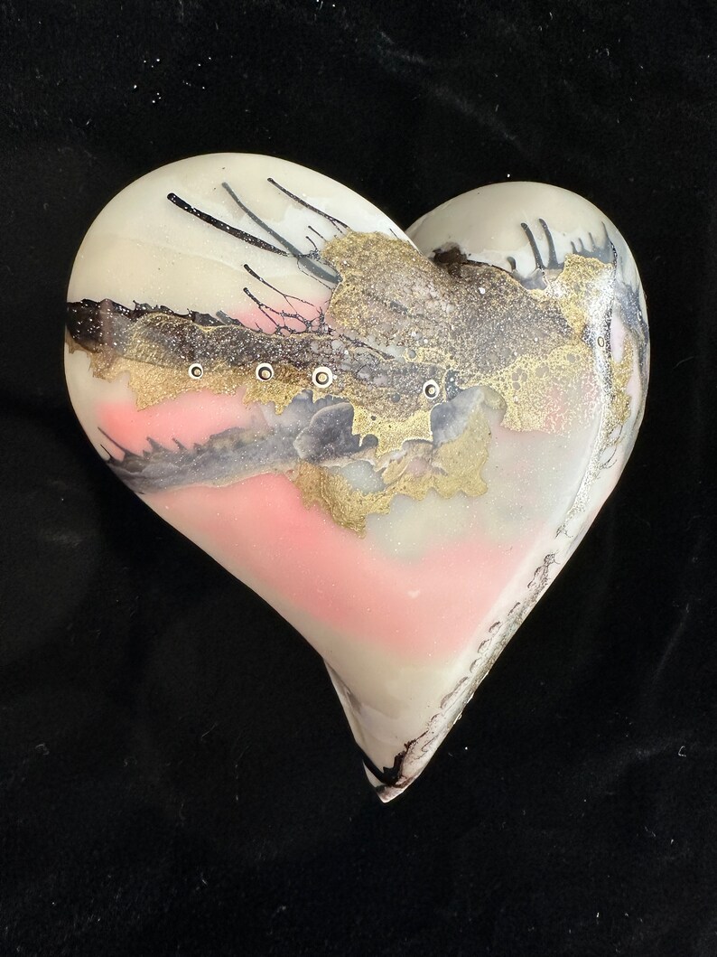 May include: A white heart-shaped glass ornament with pink, gold, and black abstract designs. The ornament has a glossy finish.