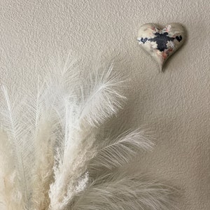 May include: A white pampas grass bouquet with a heart-shaped wall decor featuring a black and white abstract design on a beige textured wall.