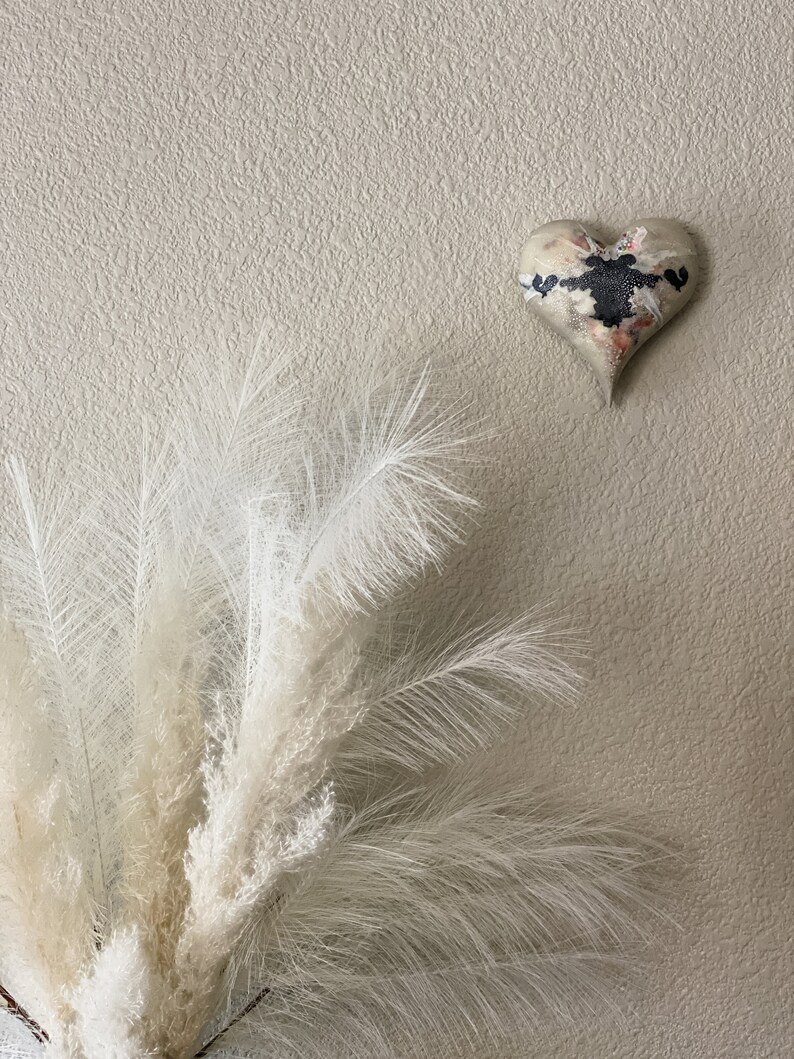 May include: A white pampas grass bouquet with a heart-shaped wall decor featuring a black and white abstract design on a beige textured wall.