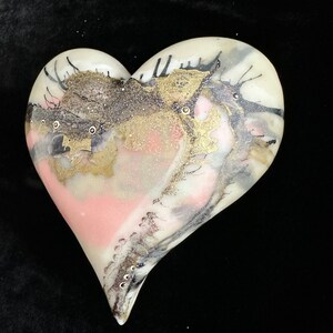 May include: A white ceramic heart with a pink, gold, and black abstract design. The heart has a glossy finish and is decorated with small, white dots.