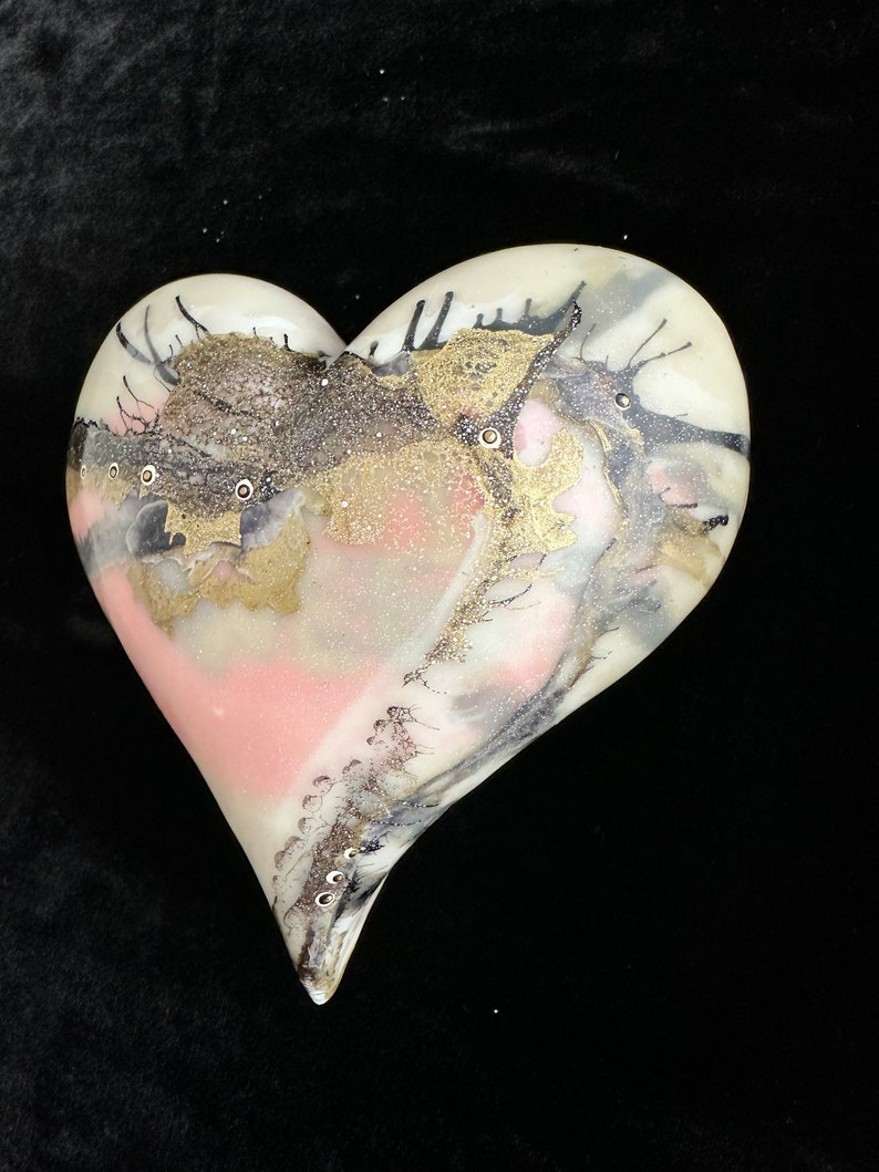 May include: A white ceramic heart with a pink, gold, and black abstract design. The heart has a glossy finish and is decorated with small, white dots.