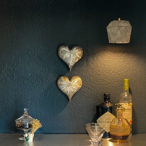 May include: Two gold heart-shaped wall decorations hang on a dark gray wall. A concrete pendant light hangs above a stainless steel countertop. A clear glass bottle of gin, a clear glass bottle of grapefruit soda, and a clear glass bottle of amber-colored liquor are on the countertop. A clear glass candy dish with a lid sits on the countertop. A small glass bowl with gold toothpicks sits on the countertop.