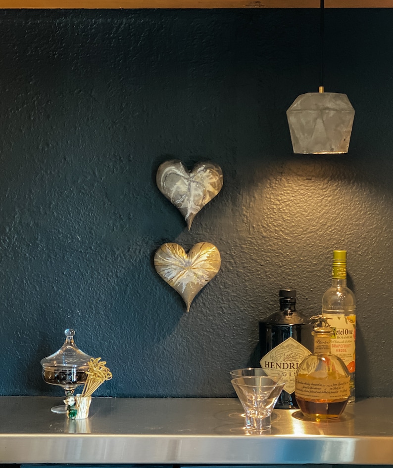 May include: Two gold heart-shaped wall decorations hang on a dark gray wall. A concrete pendant light hangs above a stainless steel countertop. A clear glass bottle of gin, a clear glass bottle of grapefruit soda, and a clear glass bottle of amber-colored liquor are on the countertop. A clear glass candy dish with a lid sits on the countertop. A small glass bowl with gold toothpicks sits on the countertop.