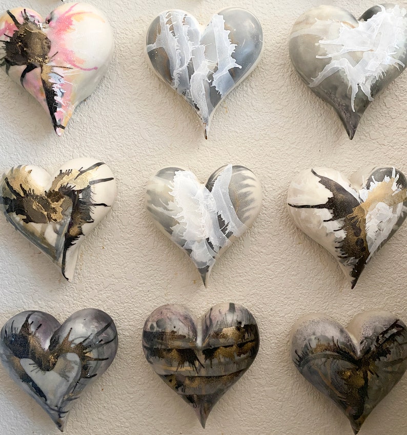 May include: Nine ceramic heart-shaped ornaments with a white base and black and gold abstract designs. The hearts are arranged in three rows of three.