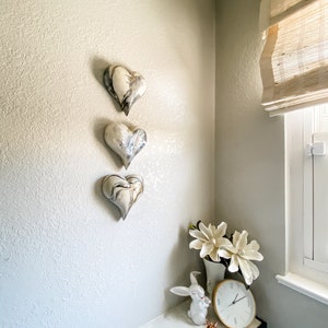 May include: Three white ceramic hearts with black and gray marbling hang on a textured wall. A white ceramic rabbit figurine and a white vase with white flowers sit on a white marble countertop.