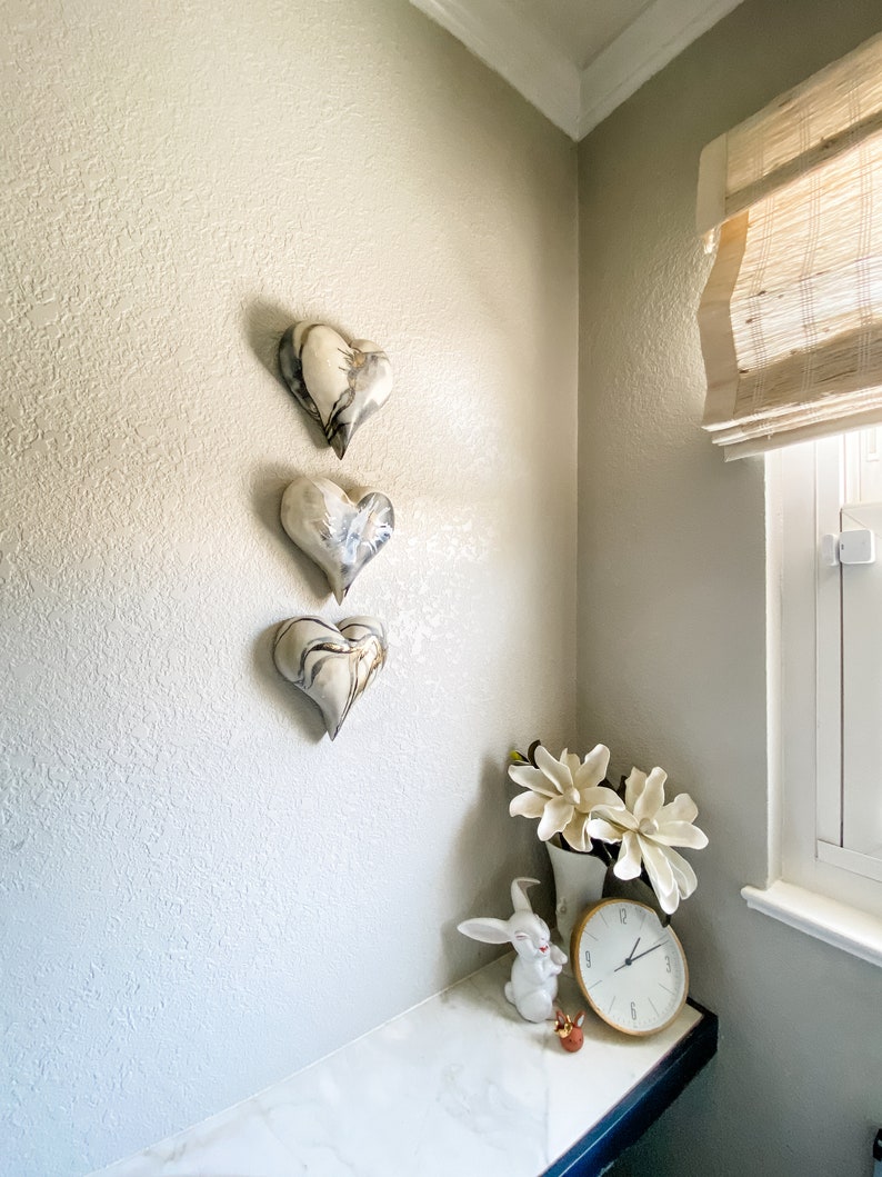 May include: Three white ceramic hearts with black and gray marbling hang on a textured wall. A white ceramic rabbit figurine and a white vase with white flowers sit on a white marble countertop.