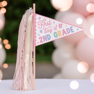 Può includere: Un gagliardetto triangolare su un bastoncino rosa con la scritta First day of 2ND GRADE in lettere colorate e piccole icone di fiori e stelle. La bandierina è decorata con una lunga nappa di lana rosa chiaro e un nastro di raso color crema.