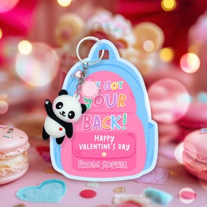 May include: A Valentine's Day themed keychain featuring a pink backpack design with the text "WE GOT YOUR BACK! HAPPY VALENTINE'S DAY FROM SOPHIA". A small panda bear charm is attached. The background includes macarons and bokeh lights.