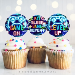 May include: Three cupcakes with white frosting and colorful sprinkles topped with round, colorful, neon-themed game controller cupcake toppers. The toppers have the text "Game On", "Eat Sleep Game Repeat", and "Level Up".