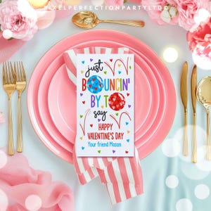 May include: A pink and white striped napkin with a white card on top. The card has a colorful illustration of bouncing balls and the text "Just Bouncing By To Say Happy Valentine's Day Your friend Mason".