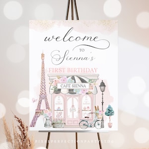 May include: A pink and white welcome sign for a first birthday party with a Parisian theme. The sign features a cartoon illustration of the Eiffel Tower, a cafe, and a bicycle with flowers. The text on the sign reads "Welcome to Sienna's First Birthday".
