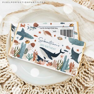 May include: A white chocolate bar wrapper with a blue and green ocean theme. The wrapper features a whale, shark, and other sea creatures. The text "Sebastian's 3rd Birthday" is printed on the wrapper.
