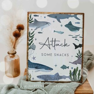 Pode incluir: Uma placa com moldura dourada, fundo branco e ilustrações azuis e verdes de tubarões, peixes e algas marinhas. O texto "Attack Some Snacks" está escrito em itálico preto.