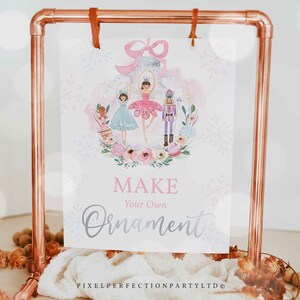 以下が含まれることがあります： バレリーナ、ナッツクラッカー、ジンジャーブレッドの女の子が描かれたピンクとシルバーのオーナメントデザインの白い看板。看板には「Make Your Own Ornament」とピンクとシルバーの文字で書かれています。