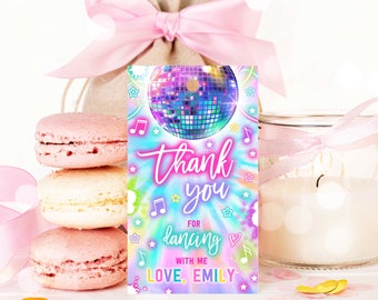 May include: A party favor tag with a disco ball graphic and the words "thank you for dancing with me, love, Emily." The tag has a tie-dye background with musical notes and stars. Pink macarons and a candle are in the background.
