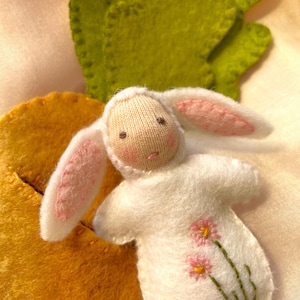 May include: A handmade white felt bunny doll with pink-lined ears and embroidered pink flowers. The doll has a knitted face and is next to a yellow felt shape and green felt leaves.