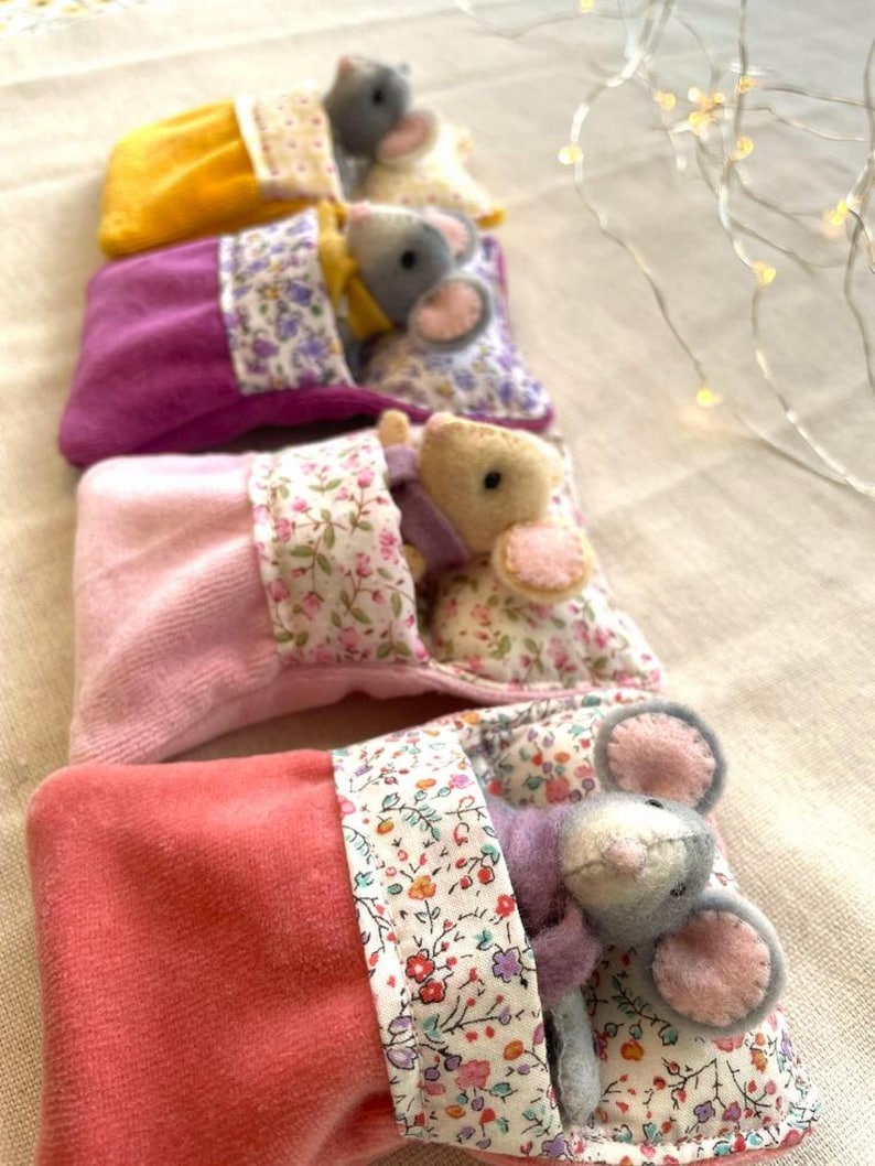 May include: A stack of handmade felt mice in colorful sleeping bags. The mice are gray with pink ears and are nestled in plush bags of yellow, purple, pink, and coral. The bags have floral patterned trim.