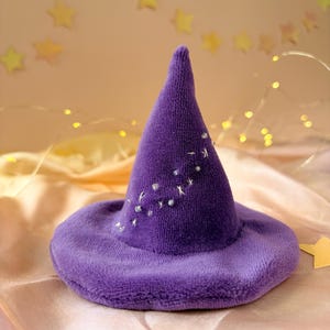 May include: A purple plush witch hat featuring a pointed crown and a wide brim. The exterior of the hat is decorated with small, scattered white embroidered star shapes and tiny clear beads that create a subtle, shimmering celestial pattern.
