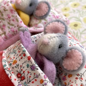 May include: Handmade felt mouse toy with gray and white fur, pink ears, and a lavender body, nestled in a floral print blanket. The blanket has a pink border and is surrounded by other similar toys, creating a cozy, whimsical scene.