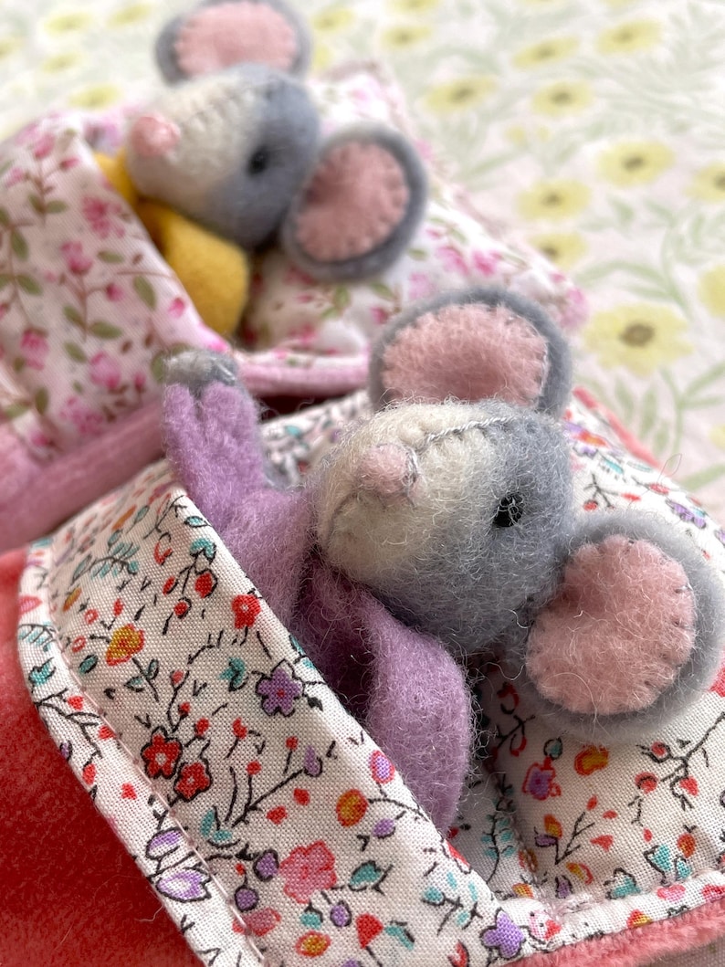 May include: Handmade felt mouse toy with gray and white fur, pink ears, and a lavender body, nestled in a floral print blanket. The blanket has a pink border and is surrounded by other similar toys, creating a cozy, whimsical scene.