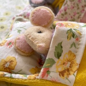 May include: A handmade stuffed animal mouse with pink felt ears peeks out from under a floral patterned blanket. The blanket has yellow and white floral patterns. The mouse is beige with a black eye.