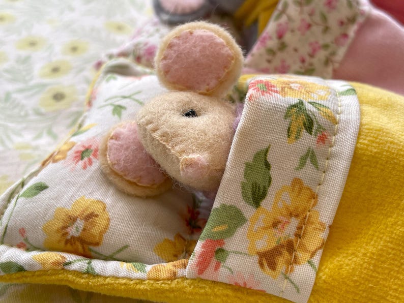 May include: A handmade stuffed animal mouse with pink felt ears peeks out from under a floral patterned blanket. The blanket has yellow and white floral patterns. The mouse is beige with a black eye.