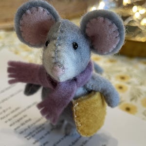 Peut inclure: Une figurine de souris en feutre gris faite à la main, avec des oreilles intérieures roses, portant une écharpe violette et tenant une sacoche jaune. La souris a des yeux en perles noires et se tient sur une feuille de papier avec du texte imprimé.