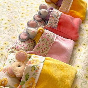 May include: A selection of handmade toy mice in colourful, plush sleeping bags and pillows. The mice have pink ears and are resting on floral patterned bedding. The sleeping bags are in shades of yellow, pink, and orange.