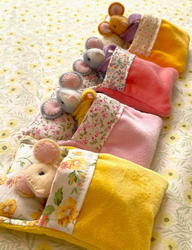 May include: A collection of handmade toy mice nestled in colorful, plush sleeping bags and pillows. The mice have pink ears and are resting on floral patterned bedding. The sleeping bags are in shades of yellow, pink, and orange.