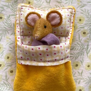 May include: A handmade yellow plush mouse toy in a yellow pocket bed. The mouse has brown fur, pink ears, and a purple blanket. The bed has a floral pattern and a white border.