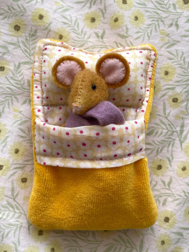 May include: A handmade yellow plush mouse toy in a yellow pocket bed. The mouse has brown fur, pink ears, and a purple blanket. The bed has a floral pattern and a white border.