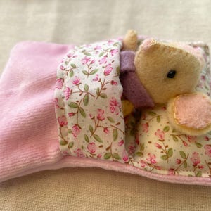 May include: A handmade plush bear nestled in a miniature bed. The bear is light yellow with a pink ear and a small black eye. The bed has a pink velvet blanket and a floral patterned pillow with pink flowers and green leaves.