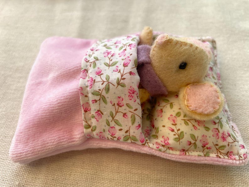 May include: A handmade plush bear nestled in a miniature bed. The bear is light yellow with a pink ear and a small black eye. The bed has a pink velvet blanket and a floral patterned pillow with pink flowers and green leaves.