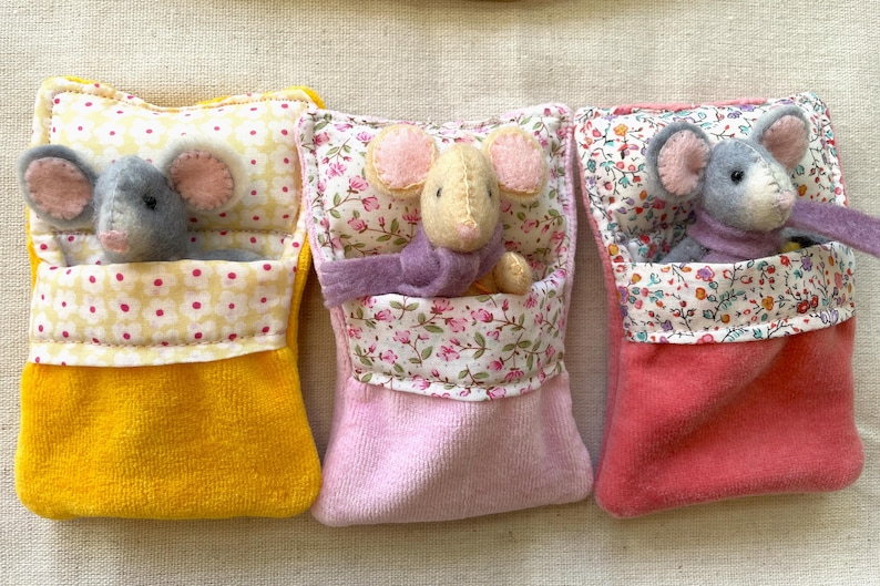 May include: Three handmade felt mouse toys nestled in colorful beds. The beds are in yellow, pink, and coral, with floral and polka dot patterns. Each mouse has large ears and a small scarf.