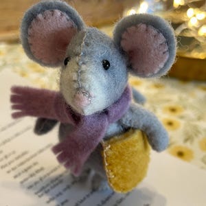 Könnte beinhalten: Eine kleine, handgefertigte Filzmausfigur in Grautönen mit großen, abgerundeten Ohren mit rosa Zentren. Die Maus trägt einen violetten Filzschal und trägt eine kleine, senfgelbe Filztasche mit sichtbaren Nähten entlang der Kanten.