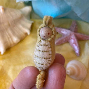 Könnte beinhalten: Eine kleine Seepferdchen-Figur aus beigem Filz mit einem hellcremefarbenen Bauch, der mit goldenen Metallic-Nähten verziert ist. Die Figur hat einen glatten, runden Holzkopf mit einfach aufgemalten Gesichtszügen und einem kleinen Kamm.