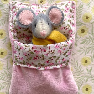 May include: A handmade plush mouse with gray fur and pink ears peeks out of a pink fabric pocket. The pocket is lined with floral fabric and a yellow scarf is around the mouse's neck. The background has a floral pattern.