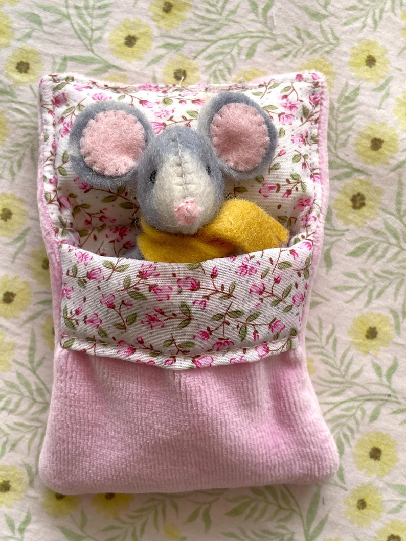 May include: A handmade plush mouse with gray fur and pink ears peeks out of a pink fabric pocket. The pocket is lined with floral fabric and a yellow scarf is around the mouse's neck. The background has a floral pattern.