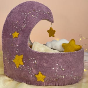 Puede incluir: Una cuna de fieltro con forma de media luna en color morado polvoriento, con enredaderas blancas bordadas, pequeñas cuentas y estrellas de fieltro amarillas. Dentro de la cuna hay una nube de fieltro blanca y un cojín de tela crema.