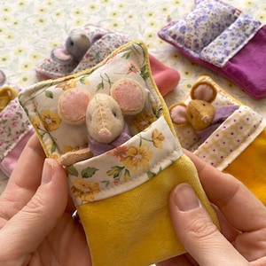 May include: Handmade felt mouse toys nestled in miniature yellow and floral-print beds. The mice have pink ears and wear purple clothing. The beds have floral and patterned fabric accents. The background is a floral pattern.