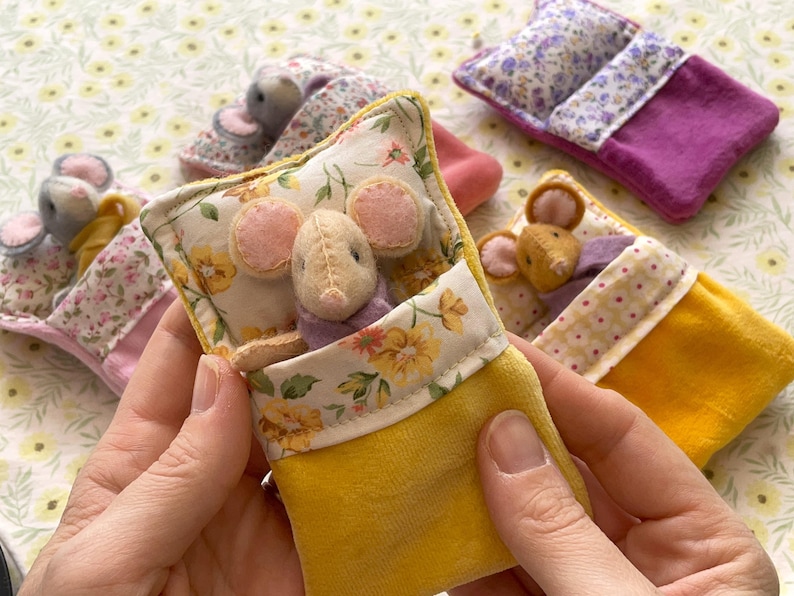 May include: Handmade felt mouse toys nestled in miniature yellow and floral-print beds. The mice have pink ears and wear purple clothing. The beds have floral and patterned fabric accents. The background is a floral pattern.