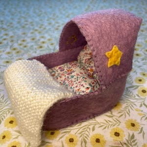 Può includere: Una culla per bambini in miniatura fatta a mano in tonalità viola, con una stella gialla. La culla presenta un cuscino a fantasia floreale e una coperta bianca lavorata a maglia, perfetta per una bambola o un oggetto decorativo.